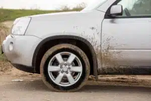 A silver car on a muddy road with mud spattered on the wheel and chassis.